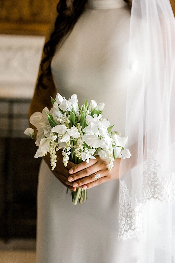 Bridal bouquet of white blooms and green foliage held by a bride in a wedding dress, with veil and engagement ring on an indoor staircase