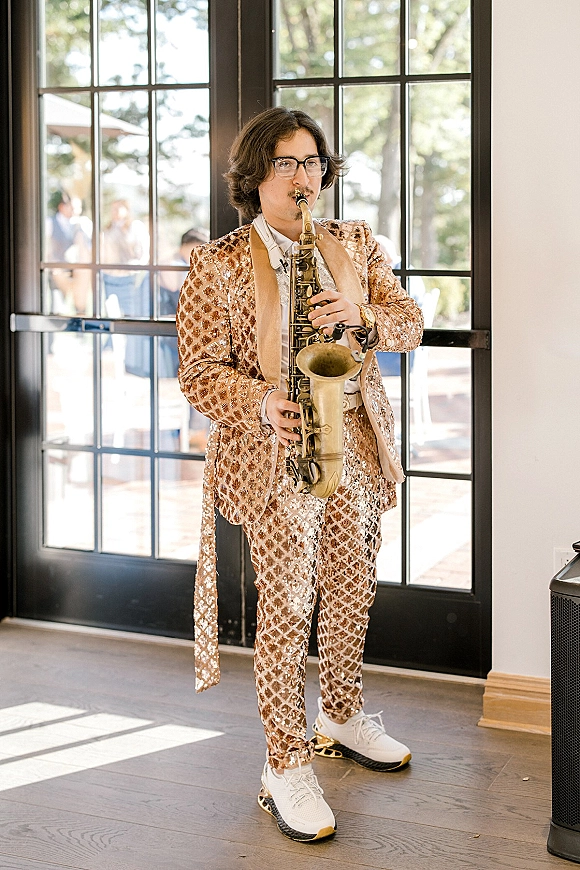 Wedding musician playing saxophone in a gold sequin suit and glasses, standing on a wood floor by large windows with guests behind