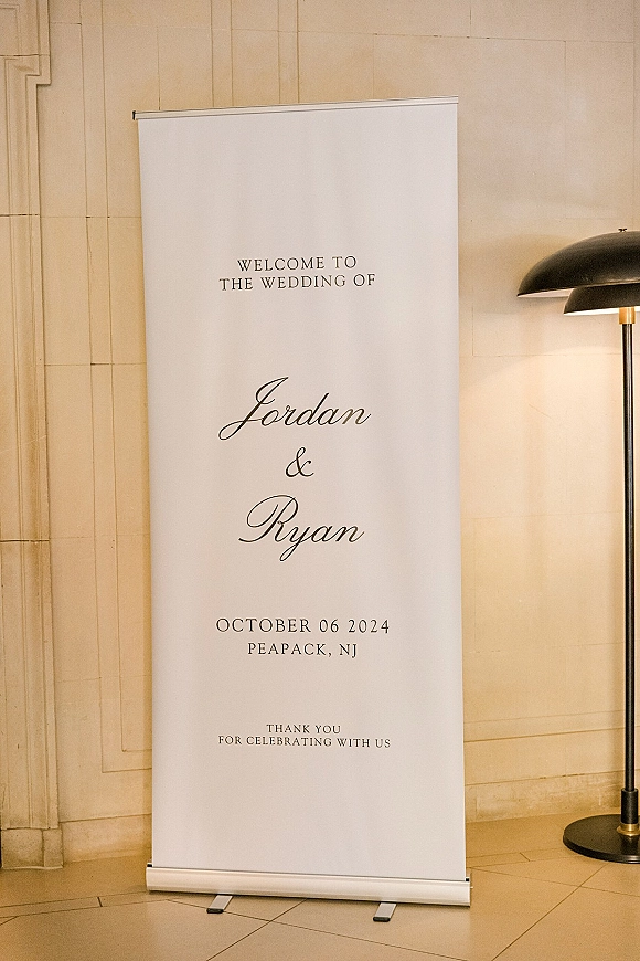 Wedding welcome sign on a freestanding banner stand, a welcome to our wedding sign beside a floor lamp against a stone wall and tiled floor