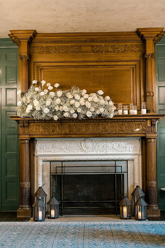 Wedding fireplace decor with mantel floral arrangement of white roses and baby's breath, pillar candles and lanterns on carved wood mantel by green walls