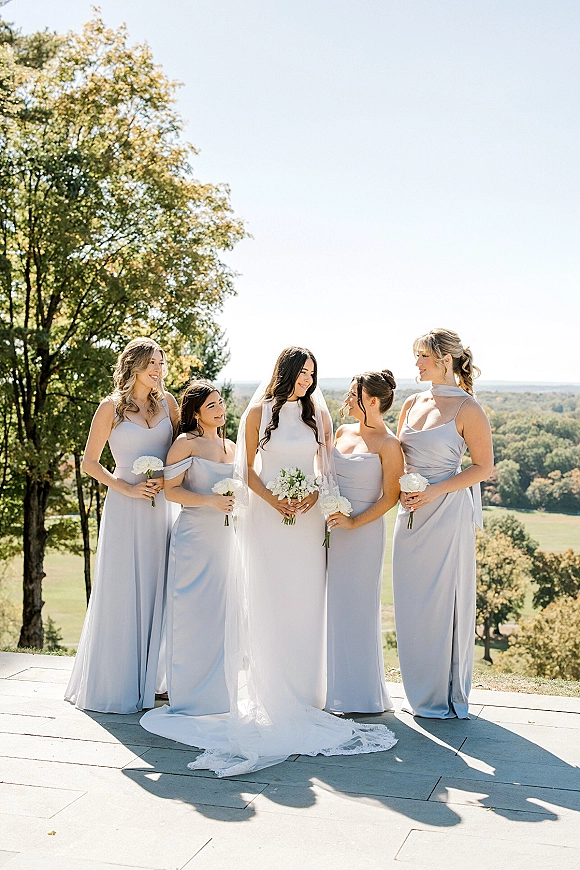 Bridesmaid group photo with bride and bridesmaids in blue dresses holding white rose bouquets on an outdoor terrace with hills beyond