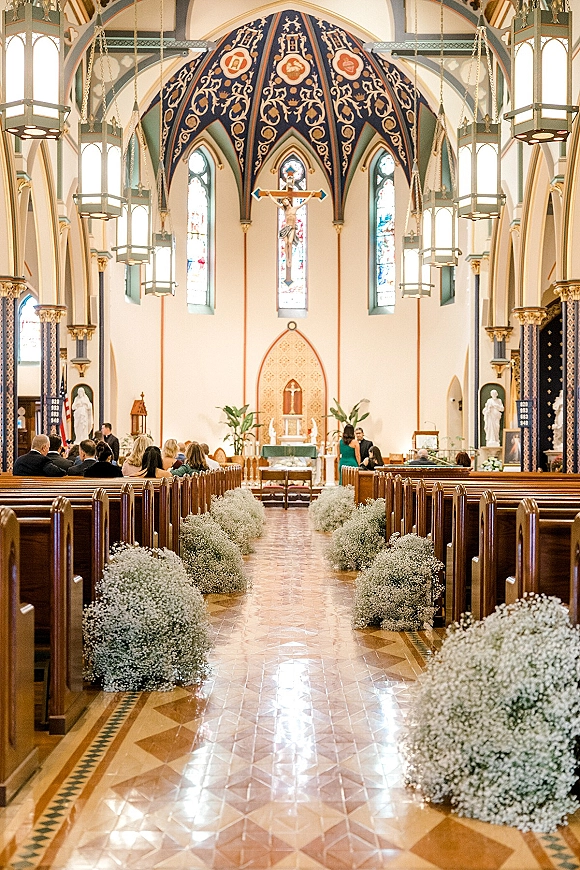 Church ceremony aisle lined with baby's breath aisle decor on wooden pews, leading to an altar with crucifix beneath stained glass windows