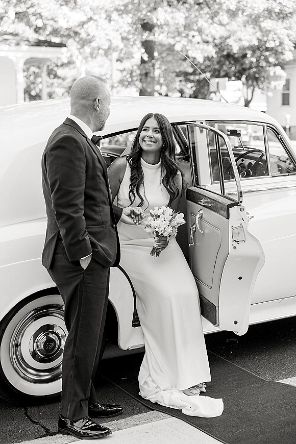 Couple portrait of bride in car as groom holds the open door beside a classic car, bouquet in hand on a quiet tree-lined street