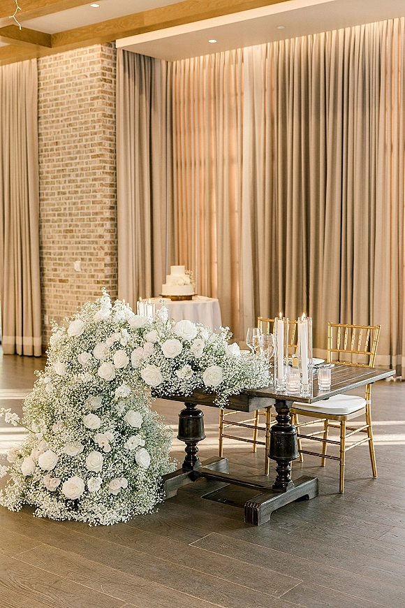 Sweetheart table decor with wedding sweetheart table flowers cascading in white roses and baby’s breath, candles and gold chairs before draped backdrop