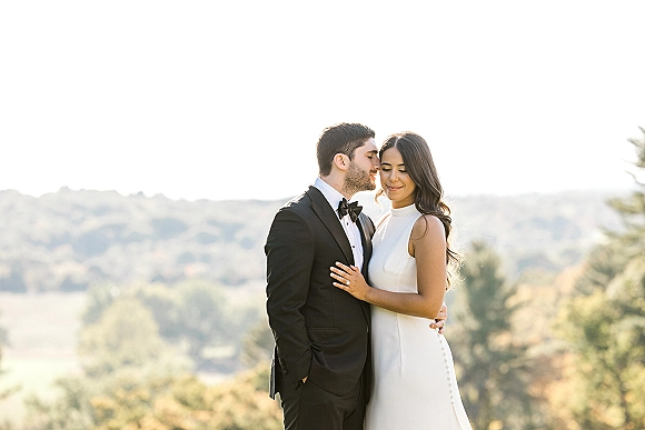 Couple portrait of bride and groom embrace as he kisses her forehead, her engagement ring visible against rolling hills and sky