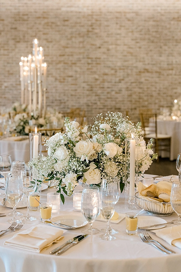 Reception tablescape with a wedding table centerpiece of white roses and baby's breath, taper candles, glassware, and brick wall backdrop