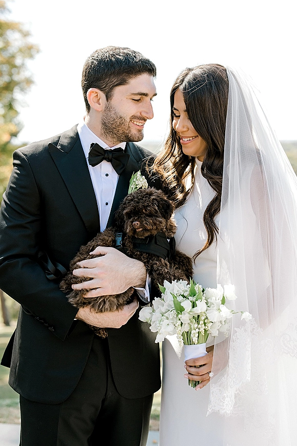 Couple portrait of bride and groom with dog, bride in long veil holding white bouquet beside groom in black tuxedo outdoors under trees