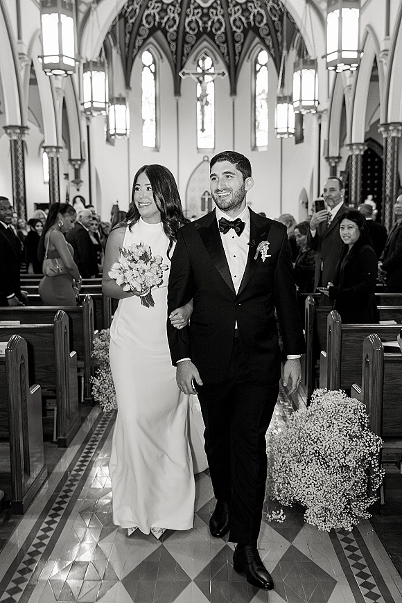 Wedding recessional bride and groom walking aisle arm in arm, bride holding bouquet, as guests watch in a lantern-lit church with stained glass.