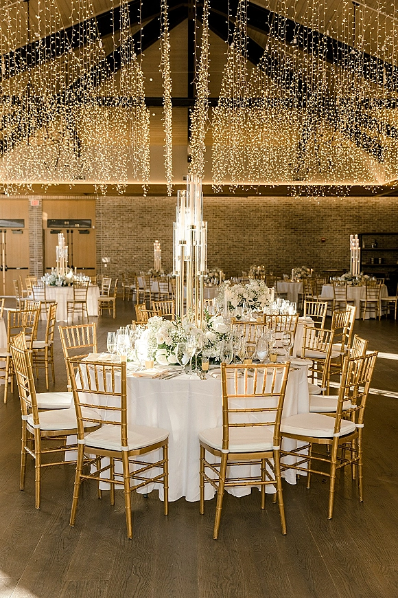 Reception tablescape with wedding string light ceiling, white floral and tall glass centerpieces, gold Chiavari chairs, and candlelit place settings in a brick-walled banquet hall