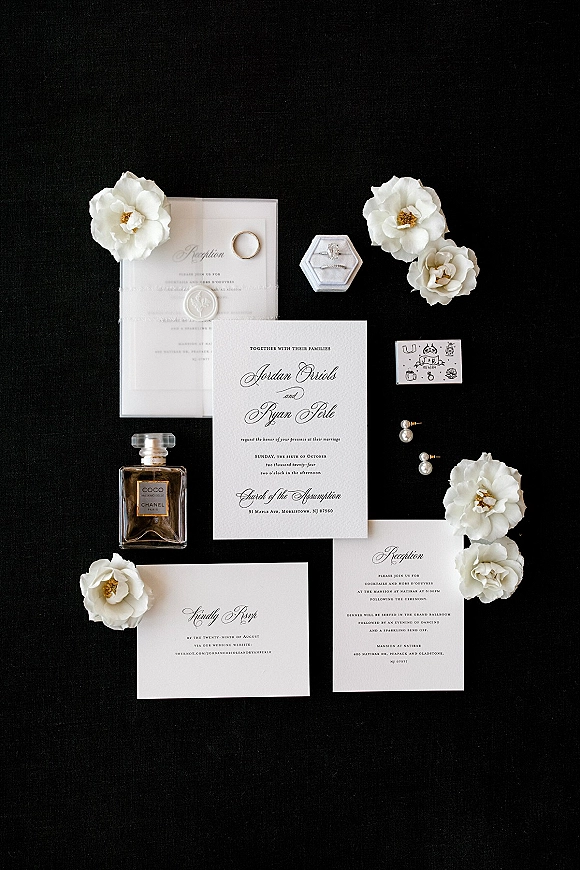 Invitation suite wedding invitation flat lay on black fabric with white flowers, wax seal, RSVP and reception cards, rings, earrings, perfume