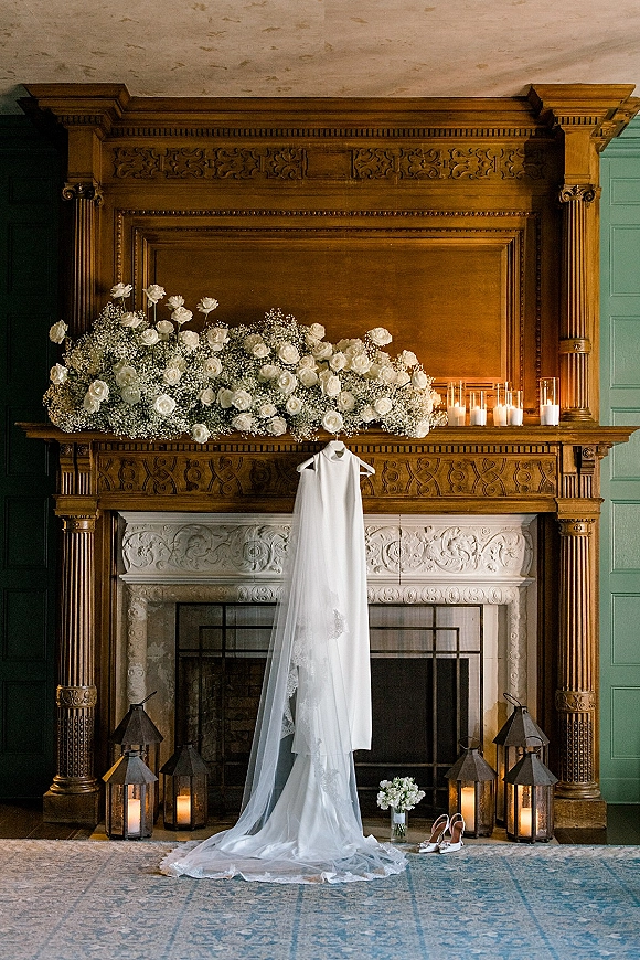 Wedding dress hanging with a lace veil, white rose bouquet and bridal shoes styled on an ornate fireplace mantel with lanterns