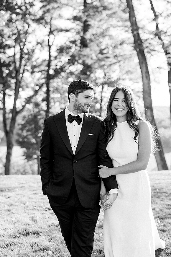 Couple portrait of bride and groom holding hands, she in a white sleeveless dress and he in a black tuxedo on a sunlit lawn