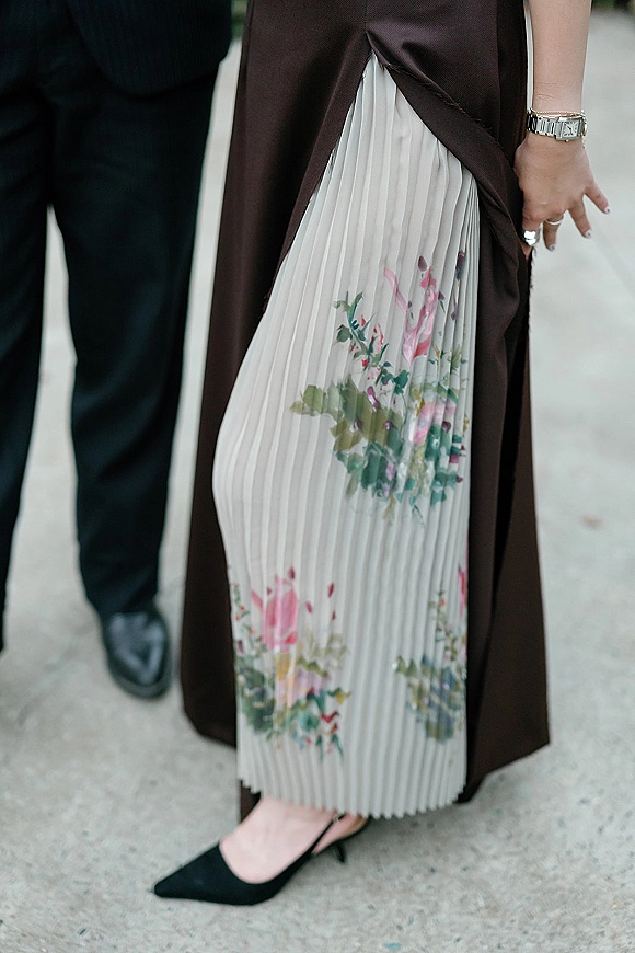 Wedding guest outfit with pleated floral midi skirt layered under a brown satin slip dress, black pointed-toe heels on a concrete sidewalk