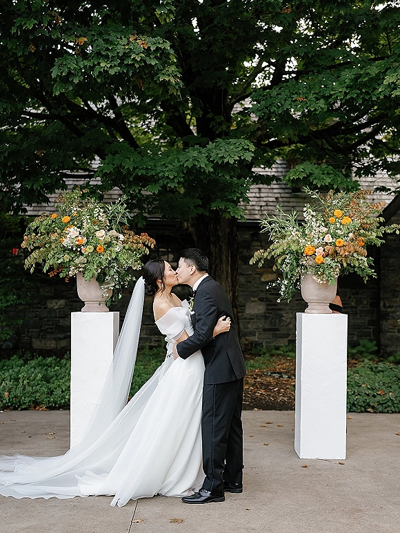 Wedding kiss between bride and groom kissing, her long veil flowing as they embrace by floral urns on a tree-lined stone walkway