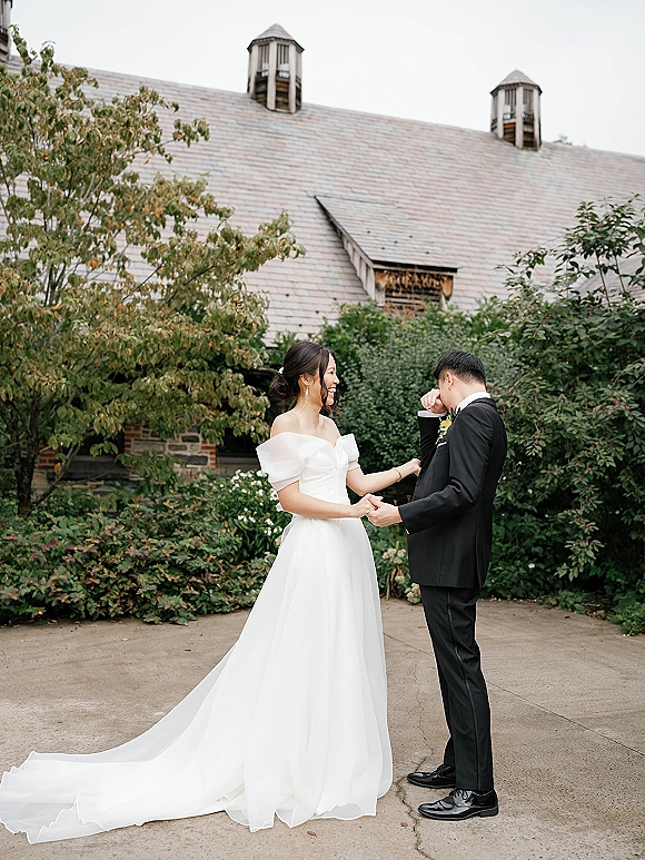 First look moment as bride in off-the-shoulder gown and long train holds groom’s hands on a walkway by shrubs and stone building