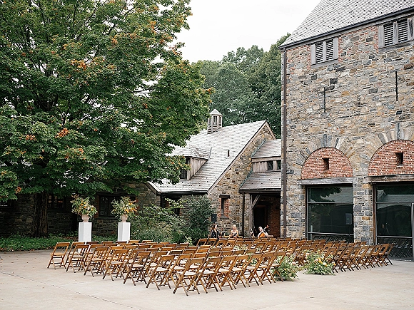 Ceremony setup in an outdoor courtyard ceremony with wood folding chair rows, white floral pedestals, and a string trio by brick arches