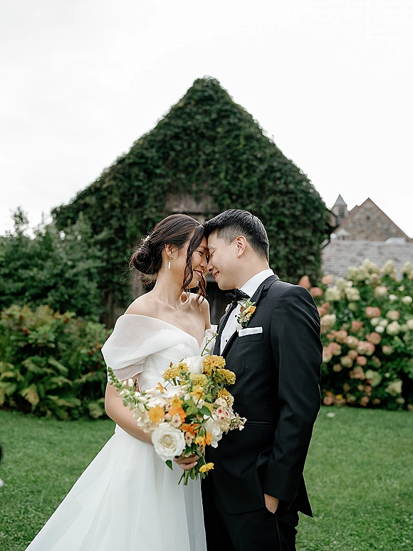 Couple portrait of bride and groom forehead touch, bride holding bouquet in off-shoulder dress before ivy-covered stone building and hedges