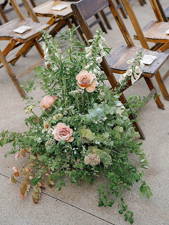 Ceremony aisle flowers in an aisle floral arrangement with blush roses, white blooms and eucalyptus beside wooden chairs on a concrete floor