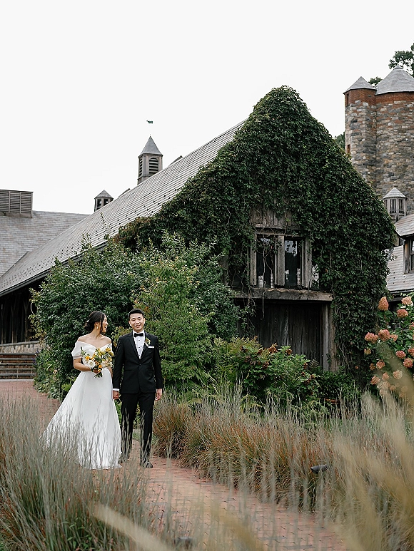 Couple portrait of bride and groom holding hands, her colorful bouquet and off-shoulder gown beside an ivy-covered stone tower on a brick path