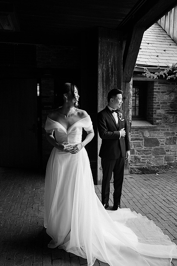 Wedding couple portrait with bride in off-the-shoulder gown and veil beside groom in tuxedo on a covered porch by a stone wall