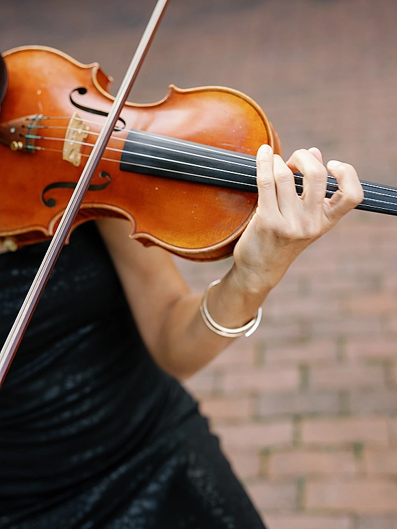 Wedding violinist in a sleeveless black dress plays at a ceremony, bowing the violin on a brick walkway with a bracelet visible