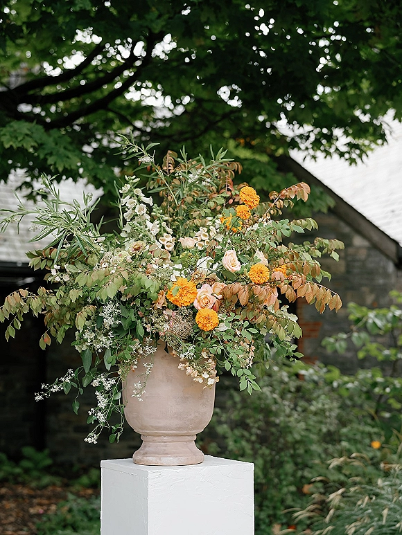 Wedding floral arrangement in an urn wedding flowers display with marigolds, blush roses, and greenery on a white pedestal in a garden setting