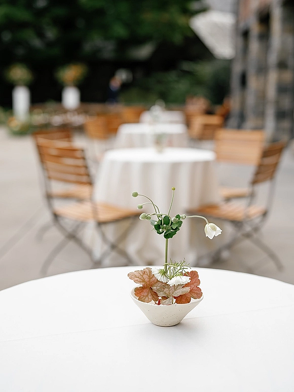 Wedding centerpieces with single stem bud vase and ceramic bowl greenery on white linens at an outdoor patio reception with wooden chairs