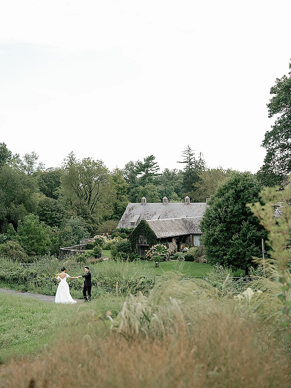 Couple portrait of bride in a strapless gown and groom in a black suit holding hands on a meadow path by a farmhouse