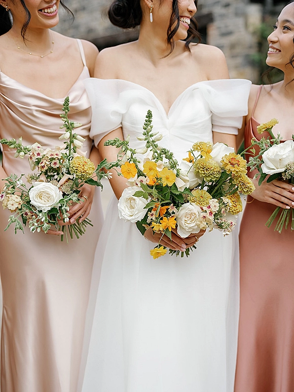 Bridesmaid portrait in satin dress holding a yellow and white bouquet with white roses and greenery against an outdoor stone wall backdrop