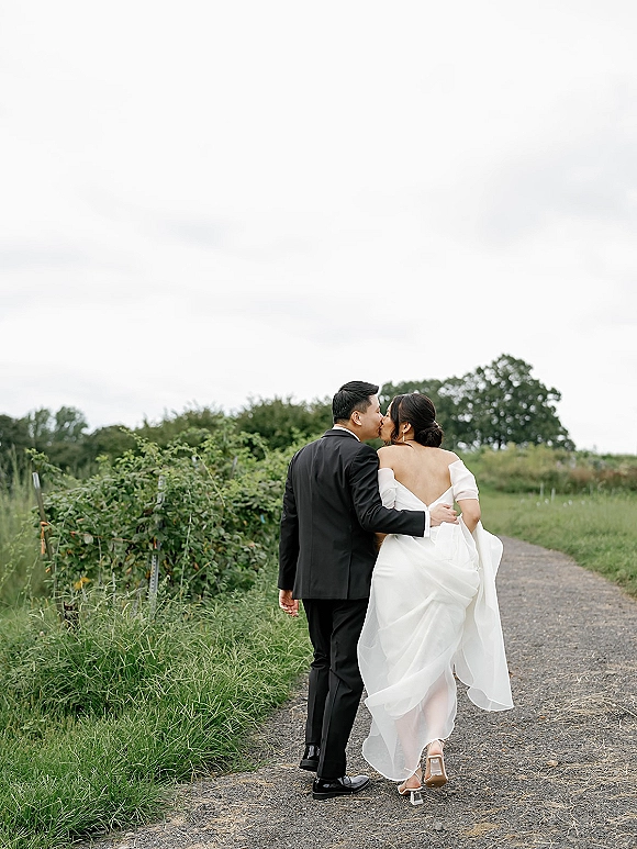 Wedding kiss portrait of bride and groom walking away hand in hand, bride in off-the-shoulder gown, on a gravel path with greenery under overcast sky