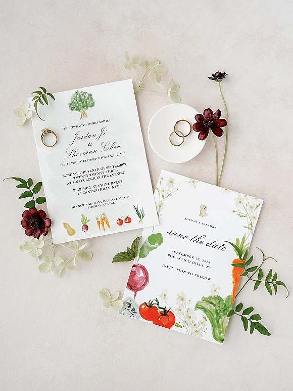 Wedding invitation suite with save the date card, calligraphy stationery, gold rings in a dish, and red floral sprigs on a neutral tabletop