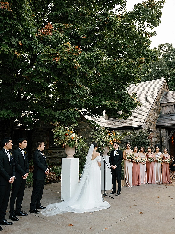Wedding ceremony with bride and groom at altar, long veil and train, bridal party in blush and tuxedos in a stone courtyard under trees