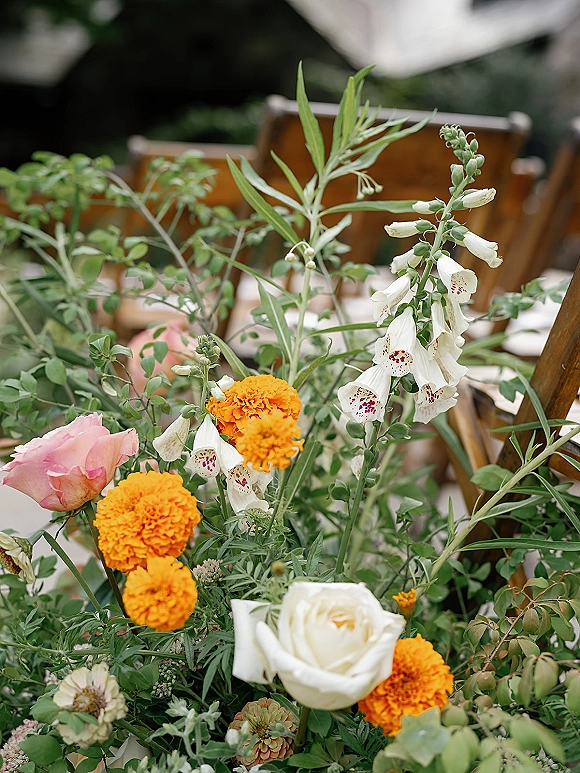 Wedding floral centerpiece with marigolds, roses, and foxglove blooms amid lush greenery on a reception table with wooden chairs behind