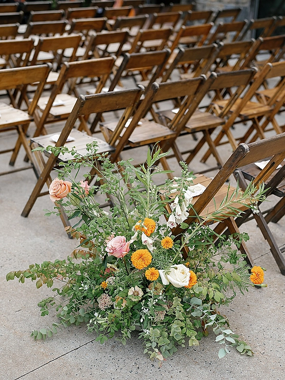 Ceremony aisle florals with grounded aisle flowers of roses and marigolds with eucalyptus foliage lining a concrete aisle beside wood chairs
