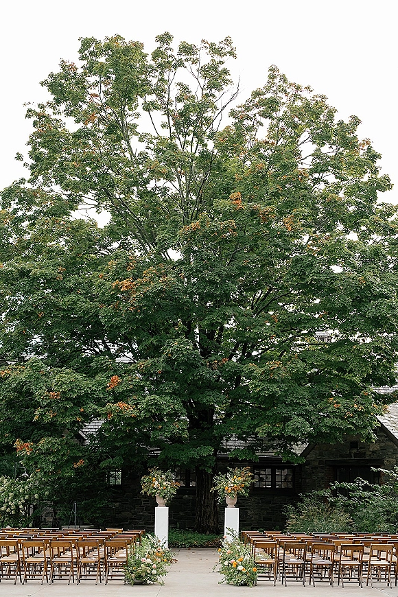Ceremony setup with wood folding chairs lining a floral aisle, framed by a large tree and stone building under an overcast sky