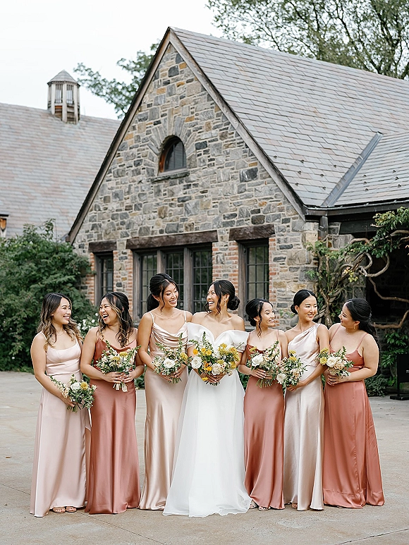 Bridesmaids portrait with bride and bridesmaids laughing, holding white and yellow bouquets in a stone courtyard with trees and windows