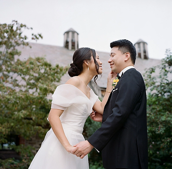 Couple portrait of bride and groom holding hands, bride touching his face, smiling in trees with roof and dormer windows behind