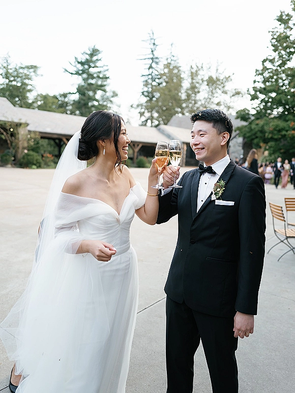 Wedding toast as bride and groom clink champagne flutes, smiling in an outdoor courtyard with mingling guests and bistro chairs