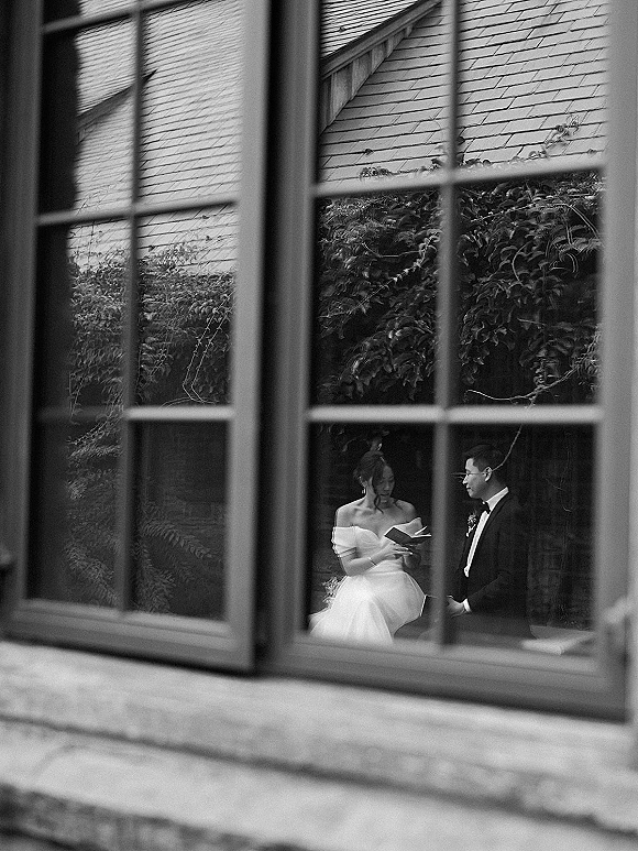 Wedding vows as bride reads from a vow book while groom listens, framed through a window with garden greenery and house exterior behind