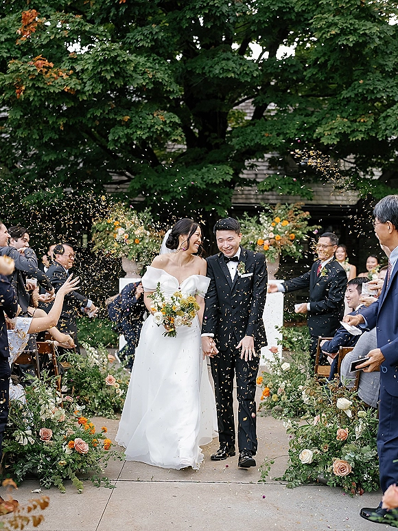 Wedding recessional with newlyweds walking aisle hand in hand as guests toss confetti, bride in off-the-shoulder gown and veil under tree canopy