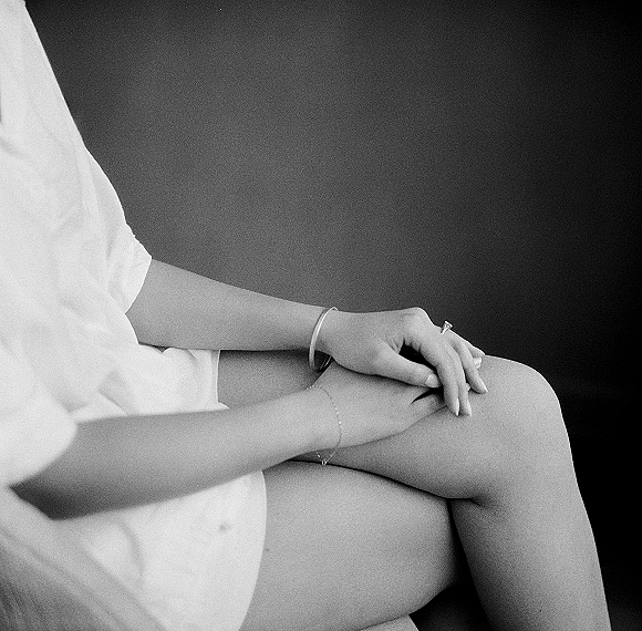 Engagement ring close-up on manicured hand, diamond ring close up with thin bangle and bracelet resting on lap in robe against dark wall