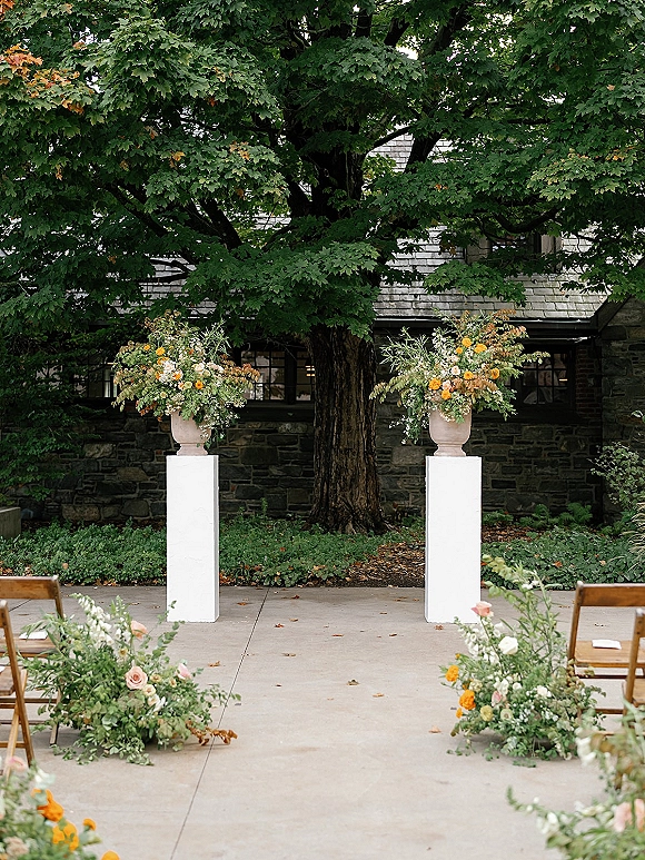 Ceremony altar decor with outdoor ceremony altar urn florals on white pedestals, aisle flowers leading to wooden chairs by a stone wall