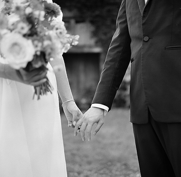 Bride and groom holding hands with a bridal bouquet, her simple wedding dress and his dark suit visible on a garden lawn