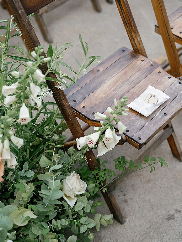Ceremony chair decor with white flowers and eucalyptus tied to a wood folding chair, plus a favor bag with sticker label on a concrete aisle
