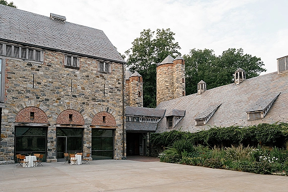 Wedding venue exterior with stone wedding venue walls, cocktail tables and wooden chairs in a courtyard with brick arches under overcast sky
