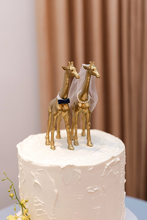 Wedding cake topper with gold giraffe figurines, mini veil and pearl necklace atop a white frosted cake against neutral drapery
