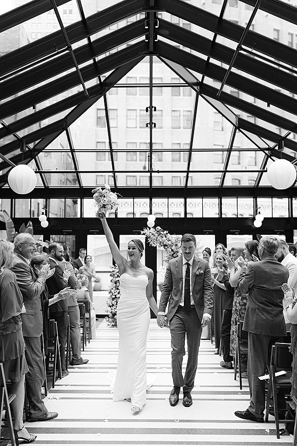 Wedding recessional as bride and groom walk down the aisle, bride raising bouquet overhead beneath a glass-ceiling atrium with cheering guests