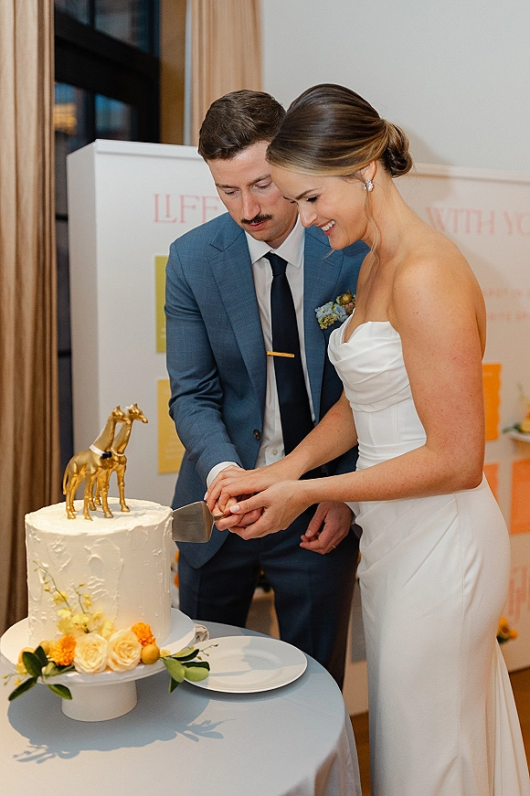 Wedding cake cutting as bride in a strapless dress and groom in a blue suit slice a buttercream cake with a gold topper at a reception table