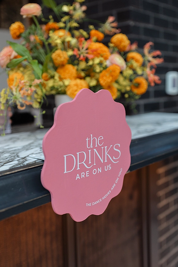 Wedding bar sign reading the drinks are on us in printed typography, with a floral arrangement in a vase on a marble bar counter