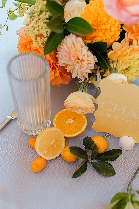 Reception tablescape with citrus wedding tablescape details, orange and yellow centerpiece in a ribbed glass vase, kumquats and place card on white linen